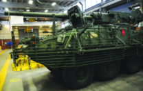 A green Stryker armored vehicle with slat armor is parked inside a maintenance bay or garage.